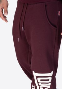 Bordeauxfarbene Sweatpants aus weichem Material mit einem Kordelzugbund, Seitentasche und großem weißen Textgrafik auf dem linken Bein.
