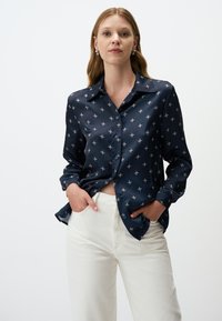 Marineblauwe zijde blouse met een subtiel bloemenpatroon, lange mouwen en een knoopsluiting aan de voorkant, gecombineerd met witte high-waisted broek.
