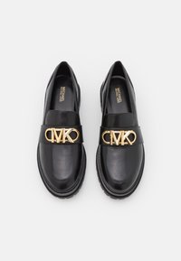 Mocassins en cuir noir avec une semelle épaisse, dotés d'un logo MK doré proéminent sur une bride élégante. Texture lisse et design à bout arrondi.