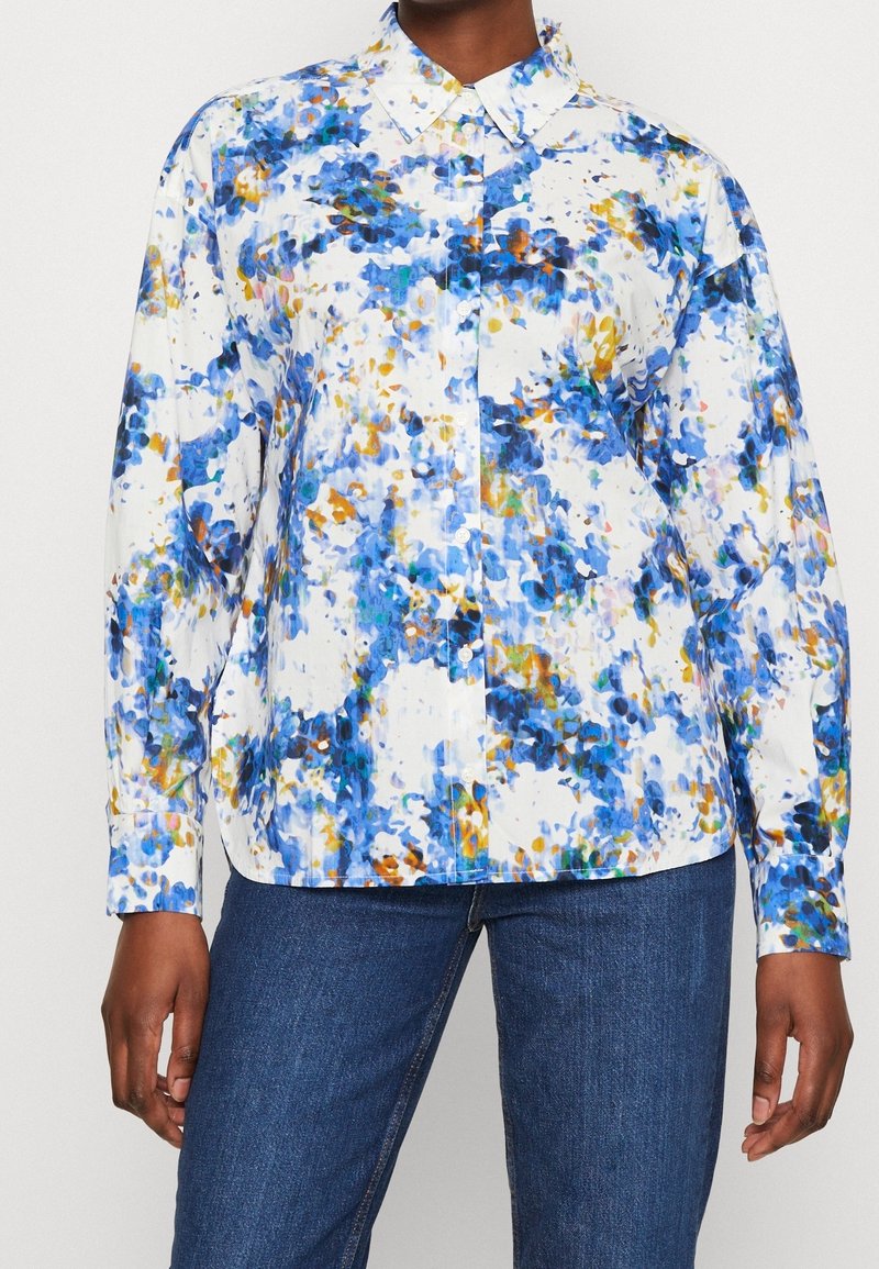 Biała koszula na guziki z niebieskim, żółtym i pomarańczowym abstrakcyjnym kwiatowym wzorem, noszona z niebieskimi dżinsami, widoczna od szyi do bioder.