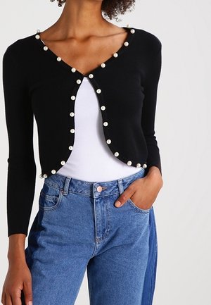 Kvinde iført en sort cropped cardigan med perleknapper over en hvid top, kombineret med højtaljede blå jeans med en hånd i lommen.