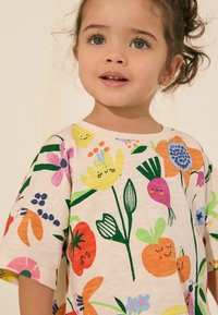 Kortærmet t-shirt med rund hals i lys beige med farverige blomster- og grøntsagsmotiver. Har et legende design og blød, bomuldsstruktur.