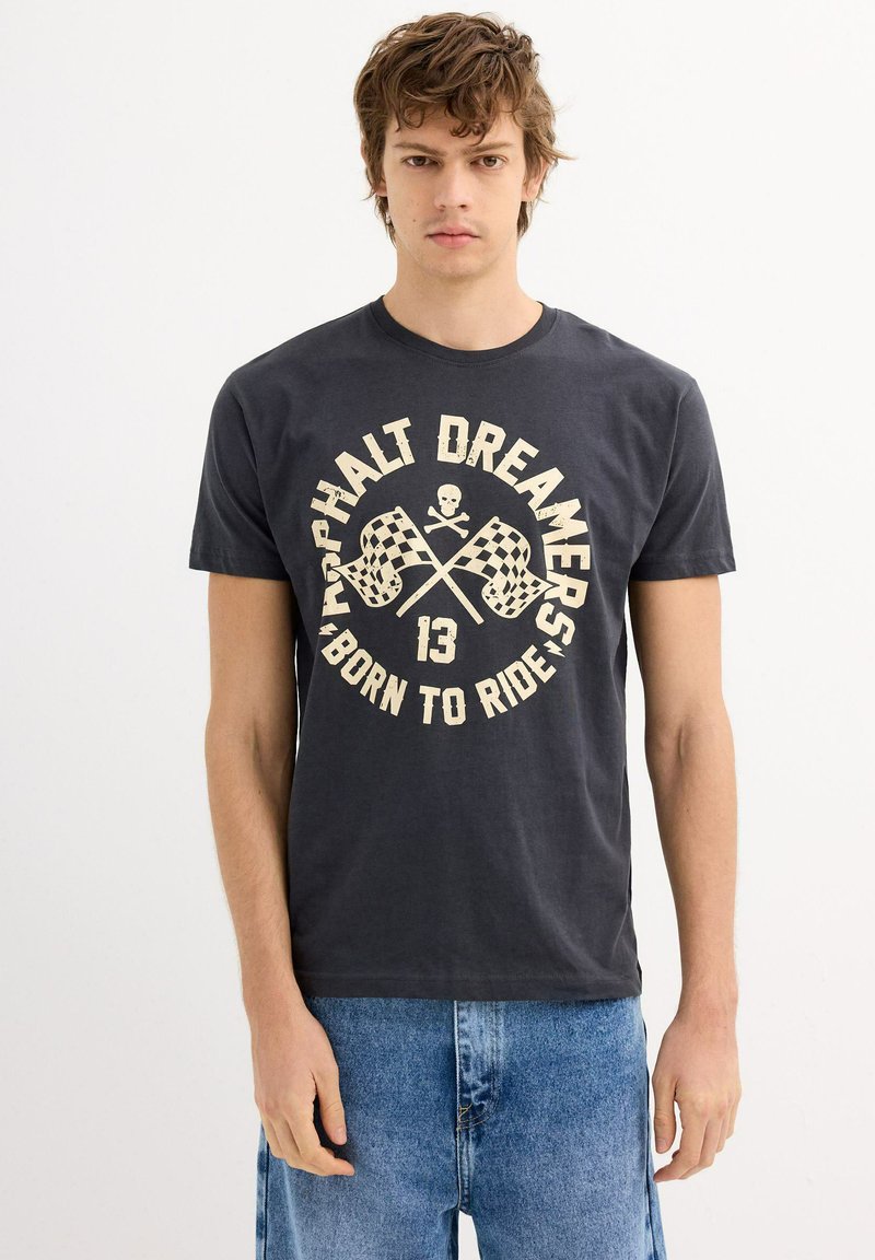 Camiseta de algodón gris con cuello redondo que presenta un diseño gráfico en crema de banderas a cuadros cruzadas y texto, combinada con pantalones cortos de mezclilla.