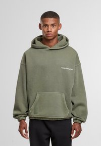 Joven vestido con una sudadera verde oliva que dice "Club de Running del Domingo", de pie contra un fondo liso gris claro.