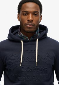 Sudadera azul marino con un logo de Superdry texturizado, cordones en color crema y una camisa de cuadros con cuello debajo.