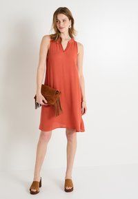 Vestido sin mangas de color óxido con escote en V, tejido texturizado y ajuste holgado, acompañado de un clutch marrón con flecos y sandalias de tacón cuadrado.
