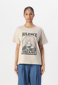 Beige bomull T-skjorte med grafisk trykk med teksten "SILENCE DREAMS", kort erm, og med løs passform. Kombinert med blå bukser.