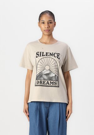 Beige bomull T-skjorte med grafisk trykk med teksten "SILENCE DREAMS", kort erm, og med løs passform. Kombinert med blå bukser.