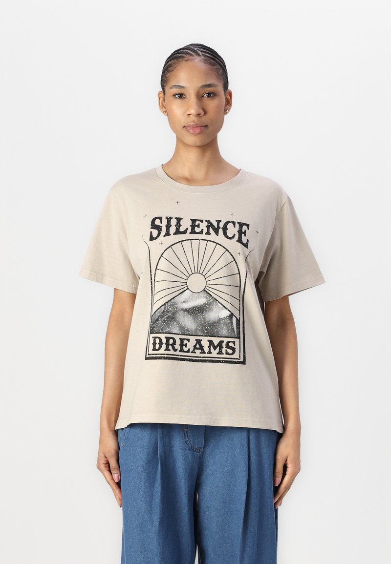 Beige bomull T-skjorte med grafisk trykk med teksten "SILENCE DREAMS", kort erm, og med løs passform. Kombinert med blå bukser.