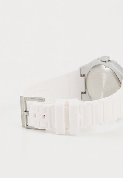 Bracelet de montre en caoutchouc blanc avec une boucle en métal argenté. Le boîtier de la montre est métallique, de forme ronde et avec des détails de cadran visibles.