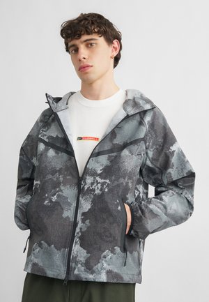 Jeune homme portant une veste zippée grise et noire à motif sur une chemise blanche avec un logo rectangulaire rouge et jaune, debout les mains dans les poches.