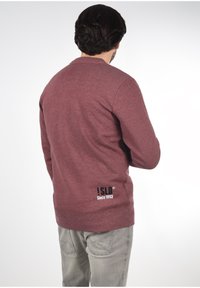 Maroon sweatshirt with a crew neck and long sleeves. Features embroidered logo text on the back at the lower left. Soft, textured fabric.