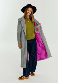 Šedý houndstooth dlouhý kabát s růžovou podšívkou, nošený přes zelený svetr, doplněný tmavými džíny a hnědými teniskami s bílými pruhy.