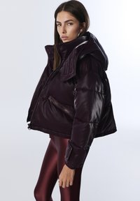Veste matelassée raccourcie en bordeaux foncé, dotée d'une finition en tissu brillant, d'une capuche oversize et de poches zippées doubles à l'avant.