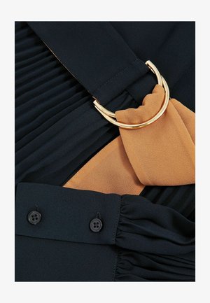 Gros plan sur un tissu noir plissé avec une écharpe beige enroulée autour d'un anneau en métal doré et une manche noire avec deux boutons.