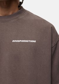 Bruine sweatshirt met ronde hals en korte mouwen, met de tekst "GOODFORNOTHING" in vette witte letters op de voorkant gedrukt.