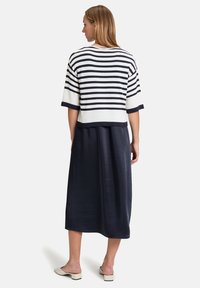 Frau, die mit dem Rücken zur Kamera steht, trägt ein weißes und marineblau gestreiftes Kurzarmoberteil, einen marineblauen Midirock und weiße Schuhe mit niedrigem Absatz.