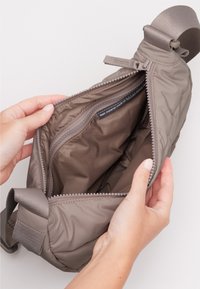 Borsa di tessuto marrone con texture, dotata di una cerniera, che mostra un interno imbottito e una tasca con cerniera, tenuta aperta da due mani. Cinturino regolabile.