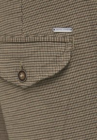 Brązowo-beżowa tkanina w kratę błędnego psa z kieszenią na klapę i ciemnym guzikiem. Srebrna metka z napisem "JACK & JONES" przyszyta do kieszeni.