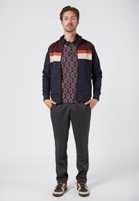 Zippered navy jacket with maroon, cream, and rust stripes; patterned shirt underneath; gray trousers; and sneakers with red accents.
