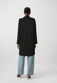Cappotto nero su misura con maniche lunghe, rever a punta e spacchi laterali, abbinato a jeans a gamba larga blu chiaro e tacchi a spillo grigi.