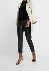 Beige trenchcoat, svart ribbad topp, svarta läderbyxor och svarta remsandaler. Outfitsen har eleganta linjer och en modern silhuett.