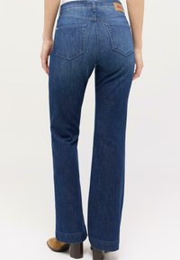 Femme portant un jean straight-leg bleu foncé avec des poches arrière et des bottes à talons beiges, se tenant contre un fond clair et uni.