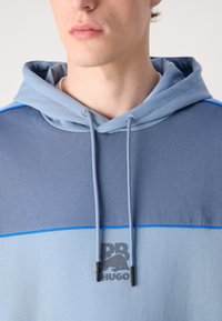 Kapučiņa kreklā zilās toņos ar priekšējo logo, kontrastējošiem krāsu paneļiem, auklām un mīkstu, teksturētu audumu.