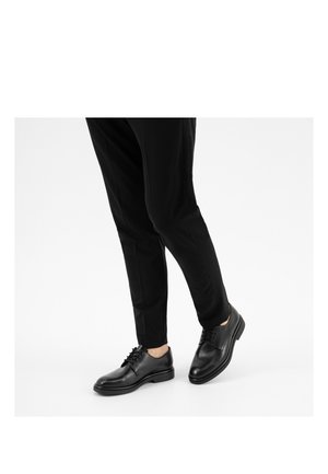 Person, die schwarze Lederschuhe und schwarze Hose trägt und einen Fuß leicht vom Boden abhebt vor weißem Hintergrund.