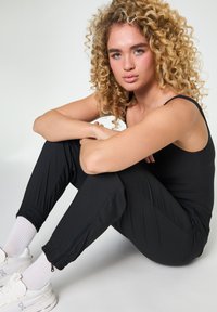 Ung kvinde med krøllet blondt hår, iført sort tanktop, sorte bukser, hvide sokker og hvide sneakers, siddende med armene viklet om knæene.