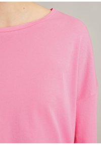 Nahaufnahme einer Person, die ein schlichtes, rosa Langarmshirt mit rundem weitem Ausschnitt vor neutralem Hintergrund trägt.