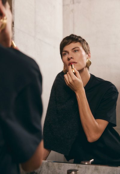 Lippenstift in einer goldenen Tube, nahe den Lippen gehalten, grauer Stoff über die Schulter drapiert, trägt ein schwarzes Shirt, hat kurzes Haar und große goldene Ohrringe.