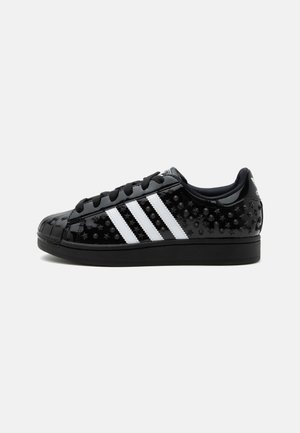 Scarpa sportiva Adidas nera con strisce bianche, con texture a motivi in rilievo a stelle e punti, e lacci neri su sfondo semplice.