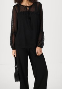 Femme portant un chemisier noir transparent à manches longues avec des pois et un pantalon noir à jambes larges, tenant un petit sac à main noir.
