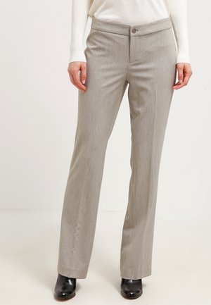 Beżowe proste spodnie o kroju taliowanym noszone z czarnymi butami i białą bluzką z długim rękawem, stojące na jednolitym jasnym tle.