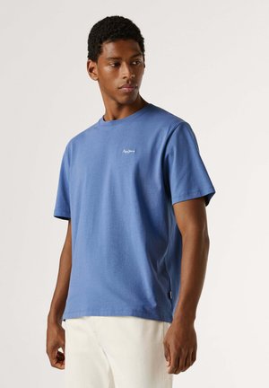 Joven con una camiseta azul de manga corta con un pequeño logotipo blanco en el pecho, combinado con pantalones color blanco roto, mirando hacia su izquierda.