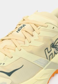 Lys cremefarvet træningssko med åndbar mesh-overdel, teksturerede detaljer, tyk polstring og grå detaljer. Har HOKA-logo på siden.