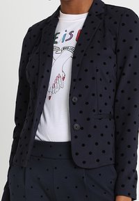 Marineblå kavaj med svarta prickar, kavajslag, två knappar och fickor, lagd över en grafisk vit t-shirt.