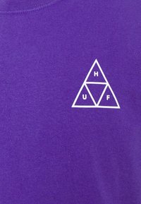 T-shirt en coton violet avec un logo triangulaire blanc portant les lettres H, U et F à l'intérieur. Couleur unie simple avec une texture lisse.