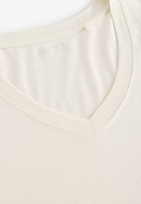 Maglietta color crema a scollo a V realizzata in tessuto morbido; presenta una cucitura delicata intorno al colletto e una texture liscia.