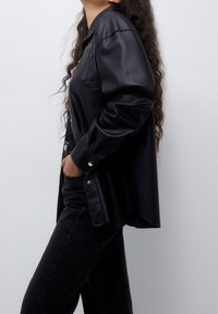 Chemise en cuir synthétique noir à coupe décontractée, manches longues, boutons pression et poche poitrine, associée à un jean en denim foncé.