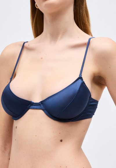 Γυναίκα που φοράει ένα σουτιέν από σατέν σε ναυτικό μπλε με μπανέλα και λεπτές ρυθμιζόμενες τιράντες, εμφανίζεται από τους ώμους έως το άνω μέρος του κορμού σε ουδέτερο φόντο.