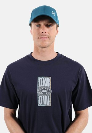 Homme portant une casquette sarcelle et un t-shirt bleu marine avec un motif central comportant les lettres OXB et OW autour d'un emblème floral.