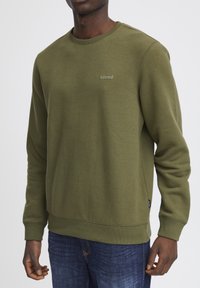Man in een olijfgroene sweater met lange mouwen en een ronde hals, met de tekst "blend" op de borst, gecombineerd met donkerblauwe spijkerbroek, staande tegen een witte achtergrond.
