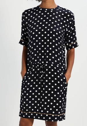 Femme portant une robe noire à manches courtes avec des pois blancs et des poches, avec une taille à cordon et une bordure dorée aux manches et à l'ourlet.