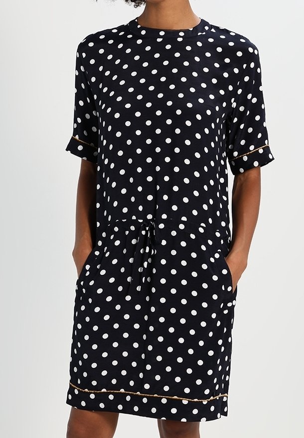 Femme portant une robe noire à manches courtes avec des pois blancs et des poches, avec une taille à cordon et une bordure dorée aux manches et à l'ourlet.