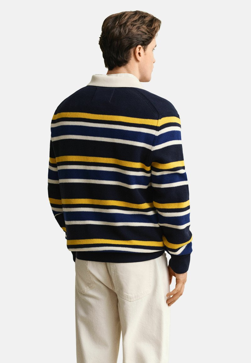 Homme de dos portant un pull bleu marine avec des rayures horizontales jaunes, blanches et bleues, et un pantalon beige clair.