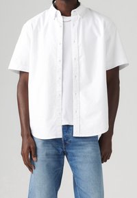 Biała koszula z krótkim rękawem, zapinana na guziki, wykonana z bawełny, z klasycznym kołnierzykiem i luźnym krojem, nałożona na białą koszulkę z okrągłym dekoltem.