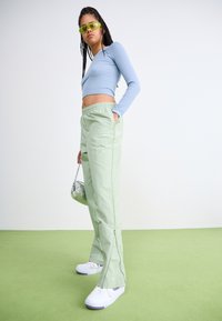 Pantalones anchos de color verde claro con cintura ajustable y cremalleras laterales, combinados con una camiseta de manga larga azul claro y zapatillas blancas.
