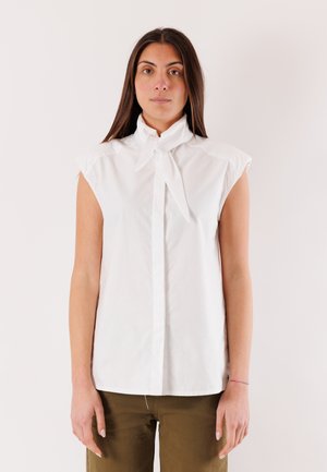 Donna con lunghi capelli scuri che indossa una camicetta bianca senza maniche con colletto annodato e pantaloni verde oliva, in piedi davanti a uno sfondo semplice.